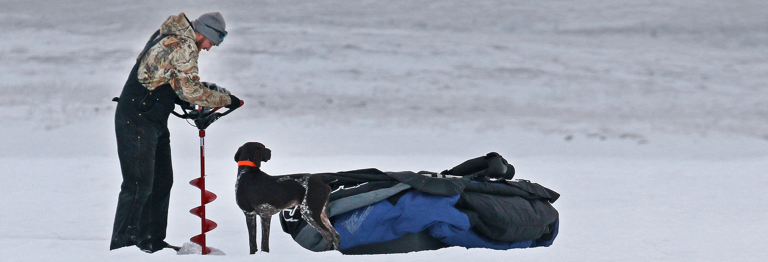 Man drilling ice hole with dog standing by