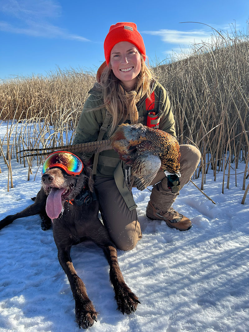 Cayla holding harvested pheasant with dog Fins lying beside her