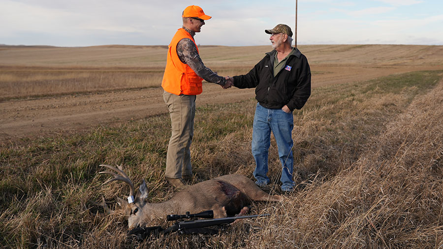 Hunter shaking hands with a landowner after a successful hunt. Harvested buck lies in front of them.