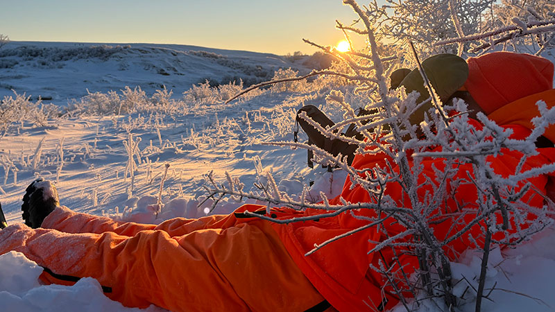 Cayla in blaze orange lying in snow looking through binoculars