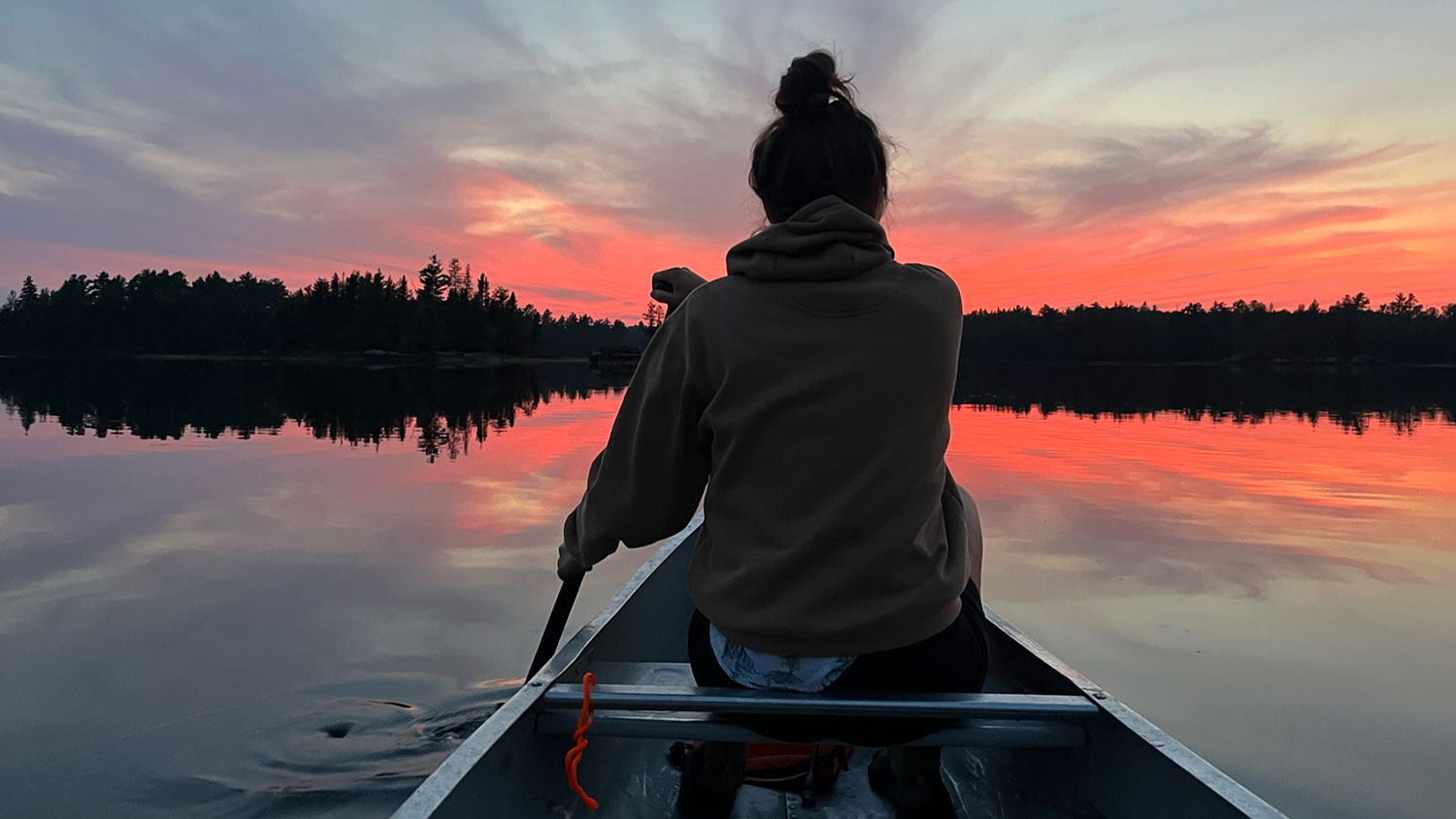 Woman paddling canoe in calm lake with sunset in front of her