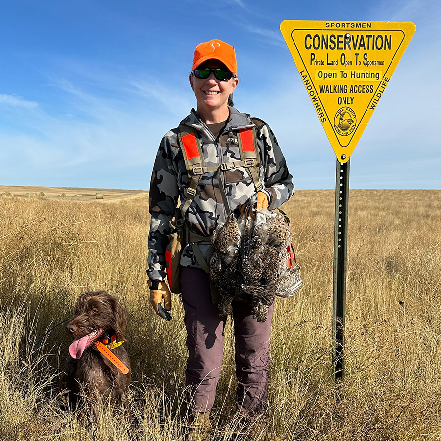 Sandy with her hunting dog and sharp-tailed grouse she shot. She is standing by a PLOTS sign.