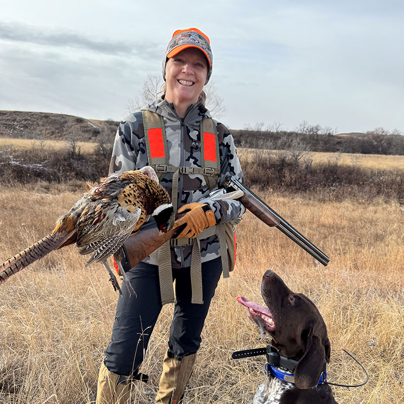 Sandy with pheasant she shot. Her hunting dog is standing by her looking up at her.