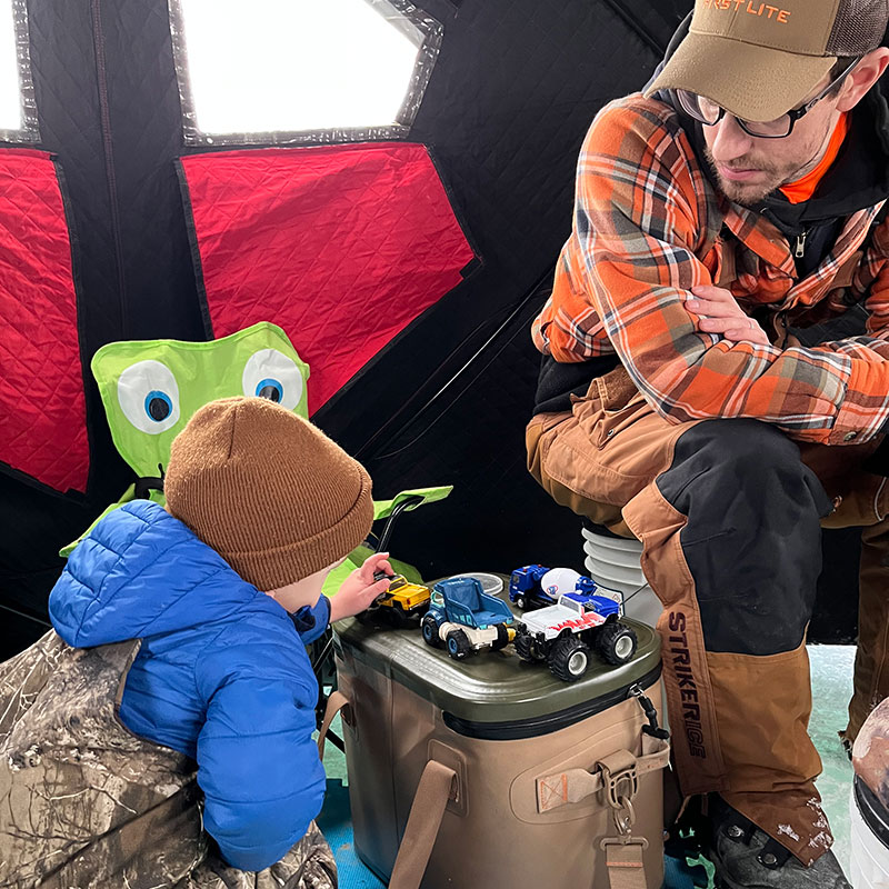 Fischer playing with toy cars in the ice house while dad sits nearby watching