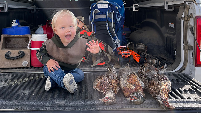 Son waving from a truck tailgate with three harvested pheasants beside him