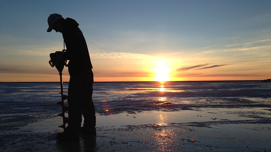 Person drilling hole in lake ice at sunrise