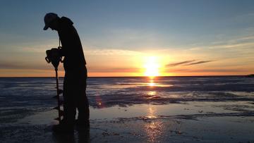 Person drilling hole in lake ice at sunrise