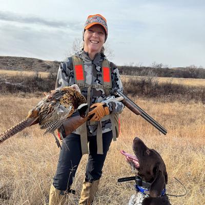 Sandra Johnson in the field holding a pheasant she shot. Her hunting dog is sitting by her looking up at her.